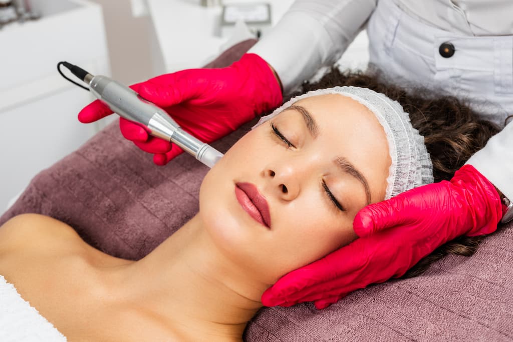 Woman receiving microneedling treatment with professional device by practitioner in red gloves, demonstrating the micro-injury creation process for collagen stimulation